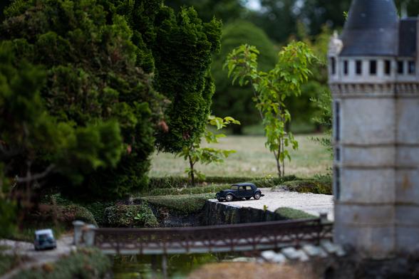 Miniature scene with toy car near a small bridge and a model of a castle turret surrounded by greenery.