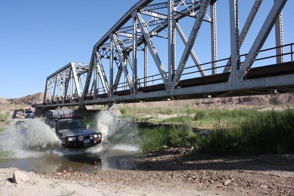 With a rail bridge looming close by, a white Toyota Land Cruiser FJ80 blasts through a stream of water on a dirt trail in the desert.