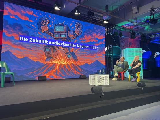 Auf der Folie ist ein "Roboter" auf einem explodierenden Vulkan und der Schriftzug "Die Zukunft audiovisueller Medien!"
