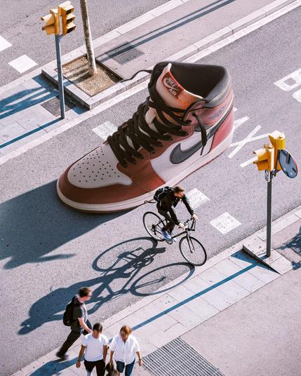 A giant Nike sneaker on a city street where pedestrians walk and a cyclist rides, creating a surreal urban scene.