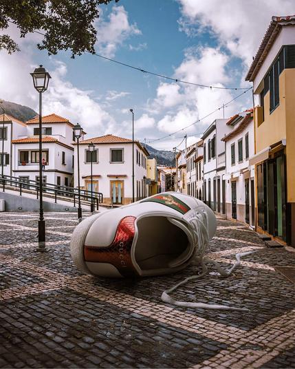 A stylish Gucci sneaker lies on cobblestone pavement, surrounded by charming white buildings under a blue sky with fluffy clouds.