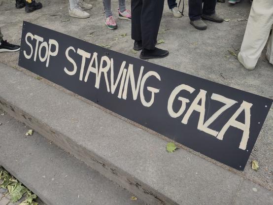 Ein schwarzes Schild steht auf dem Boden. Weiße Schrift: STOP STARVING GAZA