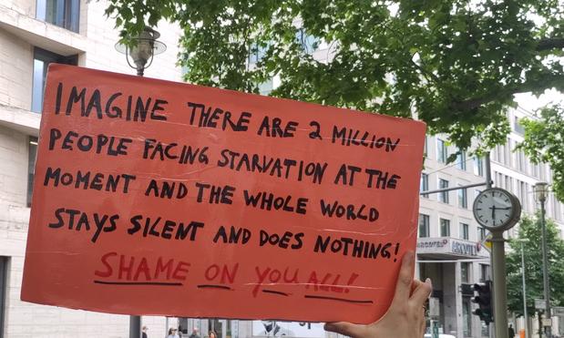 Ein rotes Plakat, mit der rechten Hand gehalten, im Hintergrund ein Baum und eine Uhr. Text: "imagine there are 2 Million people facing starvation at the moment and the whole world stays silent and does nothing." In red: "Shame on you all"