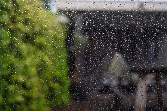 Raindrops on a window with blurred greenery and buildings in the background.