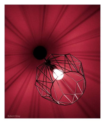 Two-tone colourised photo of a wire ceiling lamp with shadows projecting radially across the ceiling.
