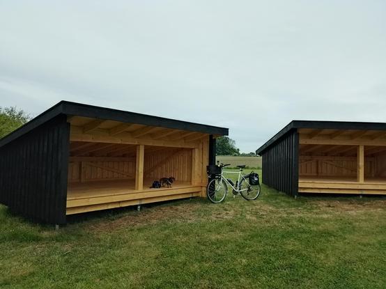 Zwei niegelnagelneue Shelter auf einer Wiese stehen nebeneinander. Es handelt sich um Übernachtungsplätze, wie sie ursprünglich von Schäfern genutzt worden sind. Die Hütten haben ein Flachdach, bestehen aus Holz und sind nach vorne offen. 
Zwischen den beiden Sheltern steht mein Rad. Effie steht in der linken Hütte und bewacht meinen Fahrradhelm.