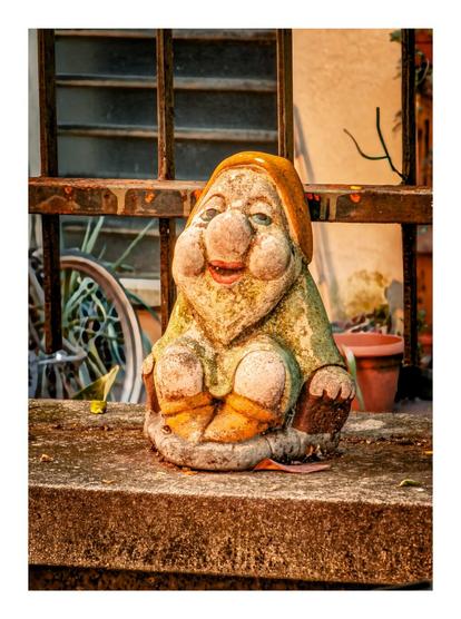 The photo is of a weathered garden gnome on a stone wall.The gnome is made of ceramic or stone and has a large nose, rosy cheeks, and a wide smile. It is wearing a green shirt and an orange hat, with its hands resting on its knees.
In the background, a rusty metal railing is visible. There is a bicycle is leaning against the wall beneath a window behind the gnome and there is also a potted plant sits to the right of the gnome.