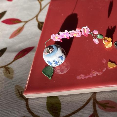 A tiny ceramic vase with blue floral patterns holds a delicate arrangement of pink orchid flowers in a single spray. The vase sits on a glossy red tile, surrounded by scattered yellow petals, and the reflection of the model can be seen in the gloss. It has a patterned fabric tablecloth in the background.