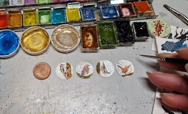 Male 20 mm winzige Miniaturen, einmal Motiv Buntspecht einmal Motive Eichelhäher einmal motivkleber und einmal Motiv Zaunkönig, dahinter die verwendeten Aquarellfarben, rechts im Bild meine Hand mit einem feinen Pinsel