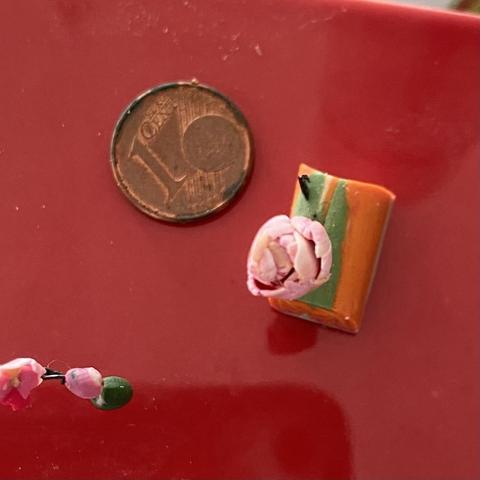 A tiny peony with overlapping pink frilly petals. It is standing in a bit of scrap clay which is orange and green. Beside it is a small 1 cent coin. In the left corner are a few of the orchid flowers from above, which also give an idea of their scale. The background to the photo is a bright red ceramic tile on which the Fimo was baked.
