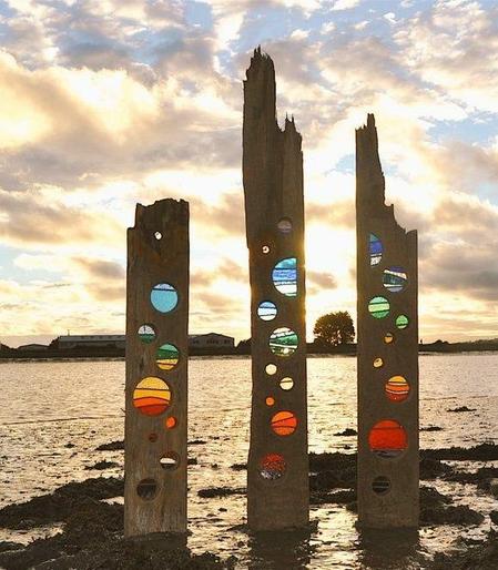 The planks of vertical driftwood placed in the sand, circles cut into them with stained glass inserted giving a rainbow bubble effect against a sunset sky