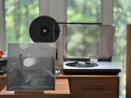 Jakob Bro - Gefion LP cover.

 A black and white photo of a man in what looks to be foul weather gear (or a parka) in a rectangular "container" looking out at the water with snow coming down. Is it a boat? It may be a boat. But it's a very rectangular boat.

The black LP plays on a vintage, restored Bang & Olufsen Beogram 4000 linear-tracking turntable to the right.