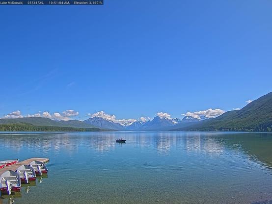 Beautiful colors in this view of Lake McDonald in Glacier National Park. Snow-capped mountains in the distance are reflected in the lake, as are the red boats in the foreground. Green trees appear on the slopes surrounding the lake. The sky is deep blue with a line of puffy white clouds on the horizon. Water clarity and sun angle combine to provide a look at the rocks on the bottom of the lake. One boat, perhaps hosting a passenger or two, appears in the center of the image.