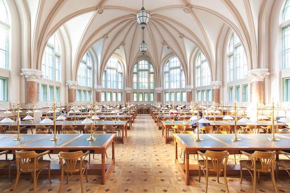 Interior of a Neogothic library reading room, showing a high rib vaulted ceiling and fan windows. Chandeliers hang from the ceiling. The room is filled with long work tables, on which a few students are seated.