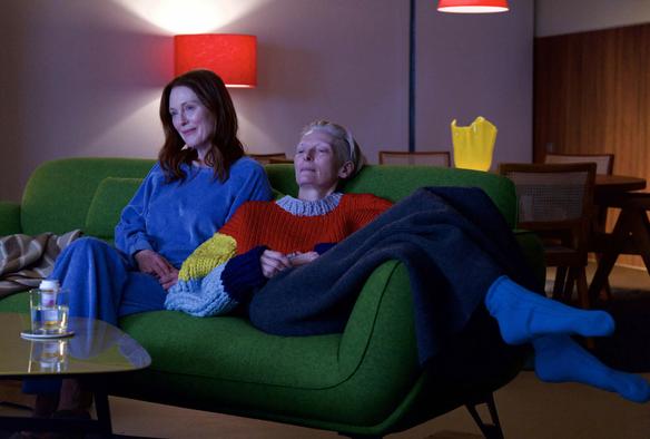 The image shows two people sitting closely on a vibrant green sofa in a dimly lit room. The person on the left is wearing a light blue outfit and has long brown hair. They sit upright with a slight smile. Next to them, the other person reclines, eyes closed, wearing a colorful sweater with large knit patterns in red, blue, yellow, and pale blue. A grey blanket covers their lap, and bright blue socks are visible. In the background, there is a table with chairs and two red lampshades emitting soft light. A yellow vase sits on the table. The atmosphere is cozy and relaxed.