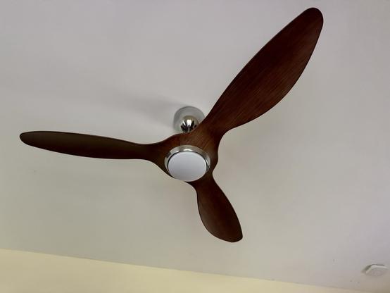 A photo of the fan installed on the ceiling. It has a shiny metal cowl to hide the electrical connections, and three brown blades. It looks a lot like an old aeroplane propeller. There’s a white circle rimmed with shiny metal at the bottom, which is the lamp.