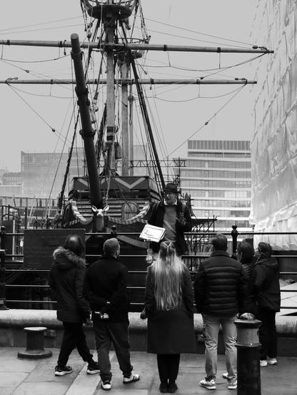 Black and white. A tour guide is talking to a group of six tourists while holding up a paper pad showing a drawing of the actual boat that's behind him - a full-sized reconstruction of the famous Elizabethan galleon The Golden Hinde, near London Bridge. Five of the tourists at the right appear to be looking at the drawing, while the rebel on the left actually appears to be looking at the boat, or rather its figurehead, a golden hind, or deer.