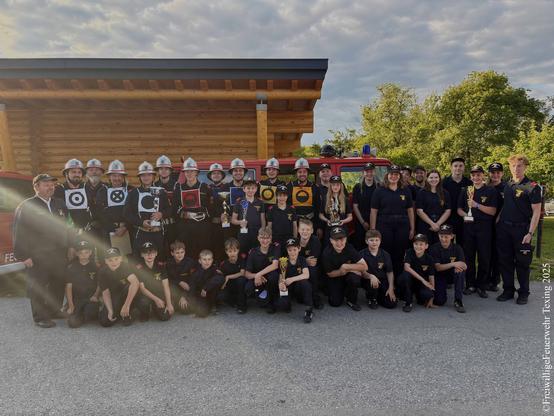 Eine große Gruppe von Kindern, Jugendlichen und Erwachsenen in Uniformen der österreichischen Feuerwehrjugend und in Uniformen der österreichischen Feuerwehren posiert mit Pokalen. 

Im Hintergrund sind eine Holzhütte und Bäume zu sehen.