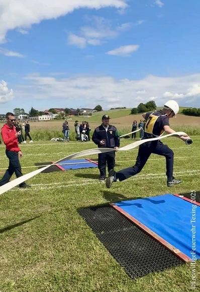 Ein Mitglied der Feuerwehrjugend springt mit einem Schlauch über einen Wassergraben. Im Hintergrund ein Bewerter in Feuerwehruniform mit Stopuhr und mehrere Zuschauer