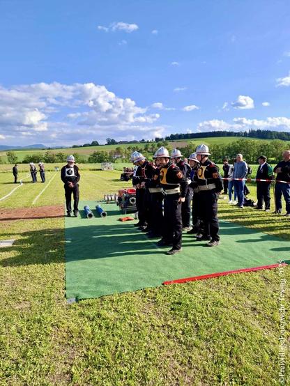 Die Bewerbsgruppe der FF Texing steht in Startformation auf einem grünen Teppich auf der Bewerbsbahn. Die Geräte sind vor der Gruppe aufgelegt, im Hintergrund sieht man auf der Nebenbahn eine Gruppe an Feuerwehrleuten mit einem Bewerter reden.