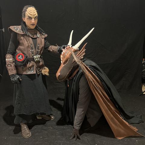 A person in a klingon cosplay stands over another person kneeling, who in a dragon cosplay with horns and wings. The background is black.