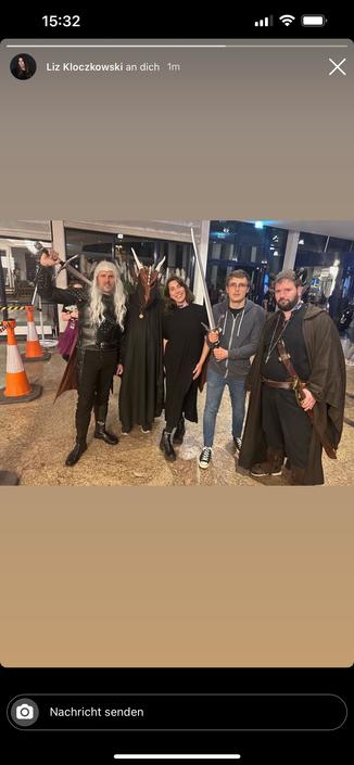 A group of five people in fantasy costumes, including capes and weapons, pose together in a brightly lit indoor setting. One person wears a mask resembling a creature, while others hold swords or are dressed in dark attire. Traffic cones are visible on the left side.