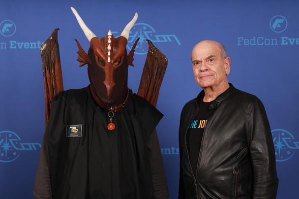 A person in a dragon costume stands next to an individual wearing a black leather jacket and a T-shirt, both posing against a blue background with event branding.