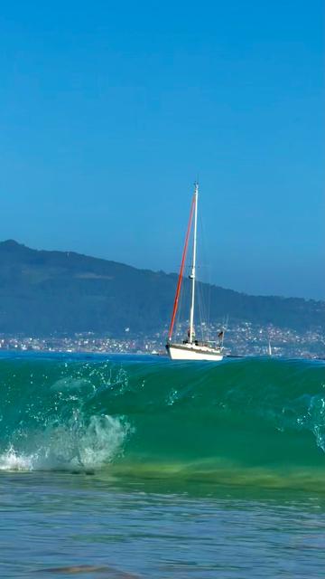 Unser Boot vom Strand aus fotografiert. Vorn bricht eine Welle, man kann in sie hinein schauen. ImmHintergrund Vigo.