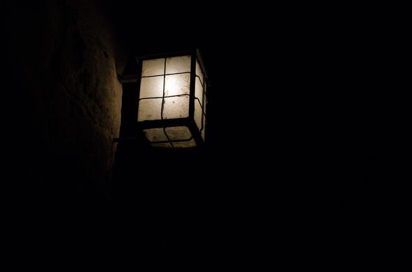 Eine rechteckige Straßenlaterne, die mit Drähten in 6 Quadrate unterteilt ist. Sie leuchtet gerade, es ist Nacht. Die Wand wird ein wenig erhellt, der Rest ist komplett im Dunkeln.