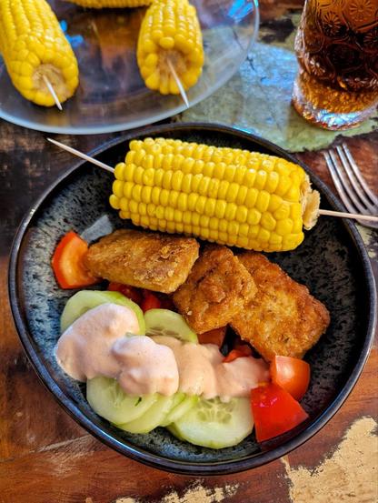 Dunkler Tisch, unregelmäßig geformte graue Schale. Darin Gurkenscheiben, Tomatenstücke mit einer rosa Sauce. Daneben panierter, braun gebratener Tofu und ein halber Maiskolben mit Zanstochern zum Festhalten. Im Hintergrund ein Teller mit Maiskolben und ein Glas mit Eistee.