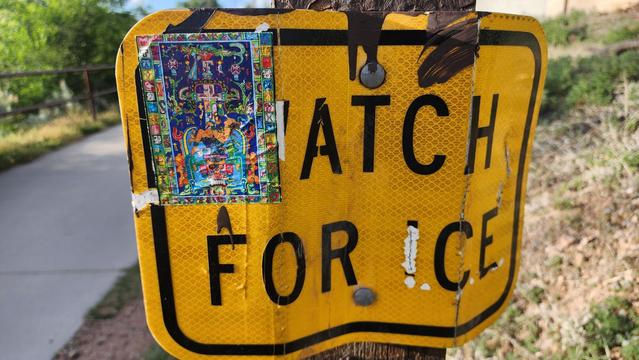 color photo of a "Watch for Ice" sign with a Mayan sticker on a riverside walk