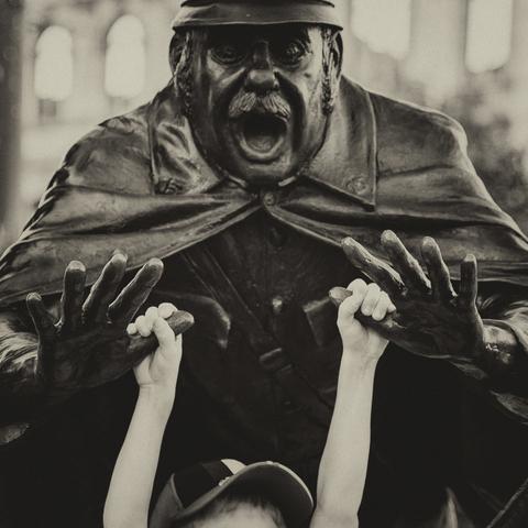Close up shot at a kid hanging from the open hands of a police man statue, clutching his metal thumbs. Only the top of the head of the kid covered by a cap, is visible as well as the top of the statue with the stunned face of the policeman and his hands pointing forward as if he was falling on the floor,  his cap is cut of the frame. The photo was processed in black and white for a timeless effect and to enhance the contrast between the white skin of the kid and the blackness of the statue.