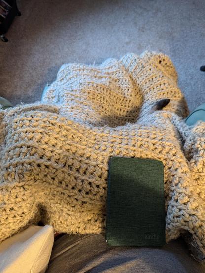 The picture is on someone’s lap covered in a blanket with a green kindle on their lap. There’s a tiny cat tail peeking out as well.