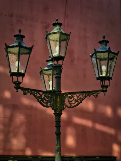 Aufnahme bei Tageslicht, im Vordergrund eine vierflammige, alte Straßenlaterne auf einem gusseisernen Laternenpfahl vor einer rostbraunroten Fassade im Hintergrund, auf die einfallende Sonnenstrahlen für einen Schattenwurf der verglasten, fünfeckigen  Lampengehäuse sorgen.