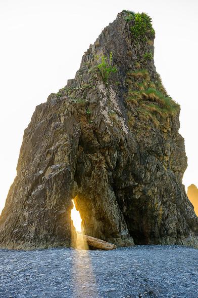 Color photo.  A tall sea stack, roughly triangular, at Ruby Beach on Washington state's Olympic Peninsula.  It rises, golden gray rock dotted with green grasses, above a blue gray rocky strip of beach.  The sky beyond it is a golden white.  A gap through the stack, at its bottom on the left, is lit golden by the setting sun hidden behind the stack.  Rays of sunlight pour through this gap, lighting a path in the rocky beach.
