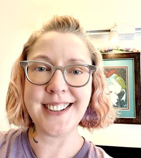 A person with short, wavy, light-colored hair wears a soft lavender shirt, the neckline slightly loose. They are wearing vintage aluminum eyeglass frames. A delicate tattoo peeks out from the side of their neck. They are positioned near a framed artwork hanging on the wall, which features a vibrant illustration of a woman with flowing hair, surrounded by intricate, swirling patterns. Above the artwork, a light fixture casts a gentle glow, accentuating the intricate lines and colors within the illustration. The mood of the scene feels contemplative and personal, blending individual style with artistic expression.