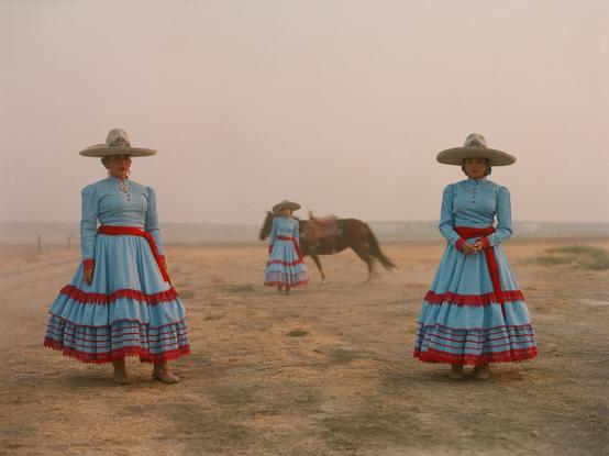 Tres mujeres jóvenes están paradas en un campo seco, con un caballo que corre atrás de ellas. Se visten trajes azul y rojo con sombreros bordados.