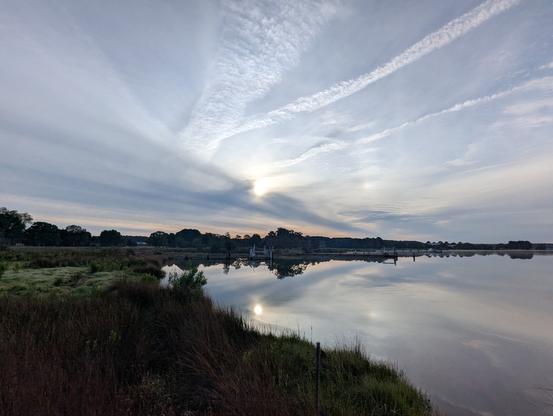 Photo of a calm morning sunrise over Harris Creek sun rising low to the left behind a gauzy grey cloud layer. A pale rainbow Sundog sits at 90° on a partial circumsolar arc. Marsh is green with riotous spring growth. Creek water is mirror flat and reflects the sun, sky and clouds.