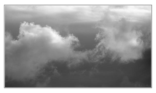 A fluffy cloud with sun brightened edges floats across the frame.  Dark moody sky below, clearer skies above.  Black and white landscape image.