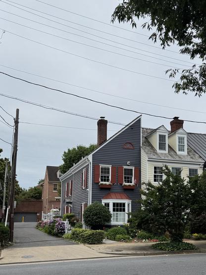The image depicts a residential street scene with two houses prominently featured. The house on the left is painted in a dark blue color with white trim and red shutters on the windows. It has a bay window with a red roof and a small arched window above it. The house on the right is cream-colored with white trim and has two dormer windows on the roof. Both houses have brick chimneys. The front yard of the blue house is adorned with a variety of plants and flowers, including purple hydrangeas. The street is lined with power lines, and there is a sidewalk visible in the foreground. The sky is overcast, and the overall atmosphere is calm and quiet.

Provided by @altbot@fuzzies.wtf, generated privately and locally using Ovis2-8B

🌱 Energy used: 0.175 Wh