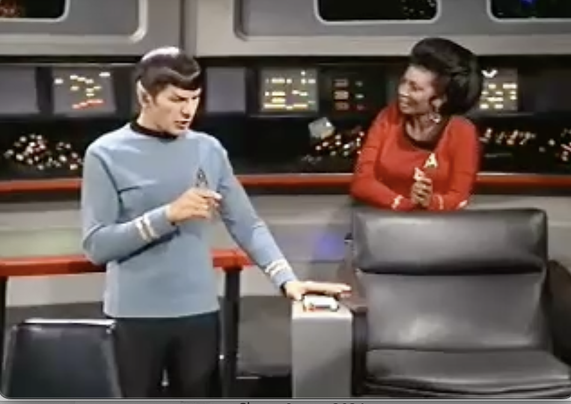 Mister Spock points at the Captain's chair of the Enterprise, explaining to a laughing Uhura that his head struck the armrest during an attack.