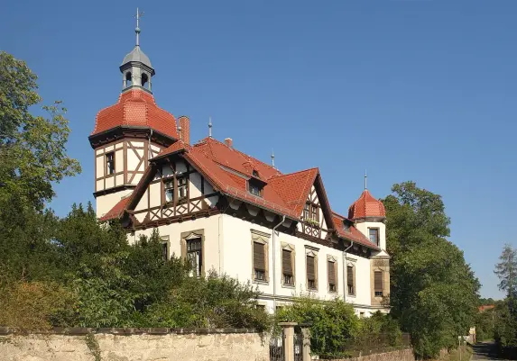 The image depicts a large, historic building that features a combination of architectural styles, likely including elements of neo-Romanesque and Gothic Revival. It has a prominent roof with distinctive chimneys and turrets, adorned with red tiles. Surrounding the structure, there are trees and greenery, contributing to a picturesque setting under a clear blue sky. The building appears to be well-maintained and may contain multiple windows with decorative wooden frames.

Image Credits: Wikimedia / User:Kolossos / CC BY-SA 3.0