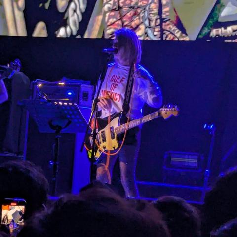 Una mujer con cabello castaño claro y corto está de pie en un escenario, tocando una guitarra eléctrica. Lleva una camiseta blanca con la frase "Golfo de México" escrita en rojo. La iluminación del escenario es azul. Hay equipo de sonido y un teclado detrás de ella. La multitud está parcialmente visible en la parte inferior de la imagen.