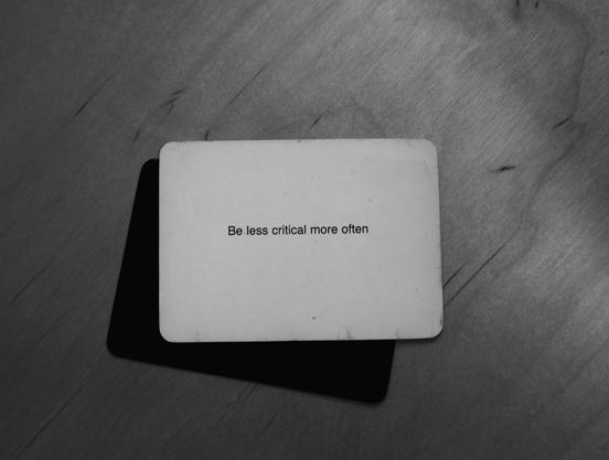 A black-and-white photo of an Oblique Strategy card. The card reads: 'Be less critical more often'.