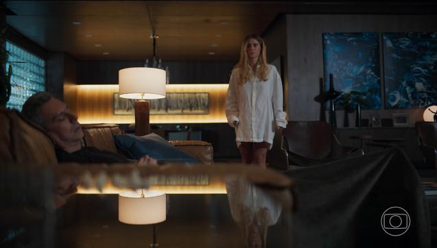 A imagem mostra uma cena da novela “Vale Tudo”, com os personagens Leila e Marco Aurélio. Marco Aurélio aparece deitado em um sofá, aparentemente adormecido ou descansando, com expressão serena. Leila está em pé, ao fundo, usando uma camisa branca longa, com uma expressão séria ou contemplativa. O ambiente é sofisticado, com iluminação indireta quente, móveis de design contemporâneo e obras de arte abstrata nas paredes. Uma luminária ao centro cria um ponto de luz suave, refletido na superfície espelhada da mesa à frente do sofá, compondo uma estética elegante e intimista. O logotipo da Rede Globo aparece no canto inferior direito.