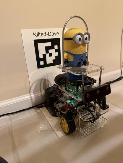 Minion Dave character atop a GoPiGo3 differential drive robot platform docked and recharging in front of an 8 by 11 printed dock sign with the robot's name "Kilted-Dave" above an ArUco Marker useful to find the dock after wandering.