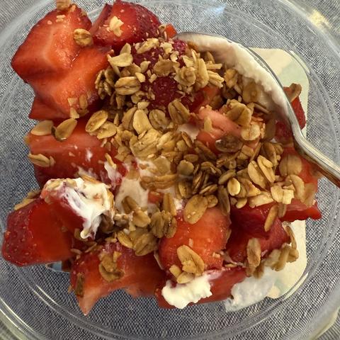 A glass bowl containing lots of ripe strawberries cut up and piled on top of yogurt and topped with a hearty sprinkle of granola.