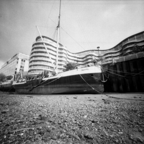 Photograph taken from the shingle/mud bank of The Thames at low tide, looking at a beaches boat with buildings in the background.
