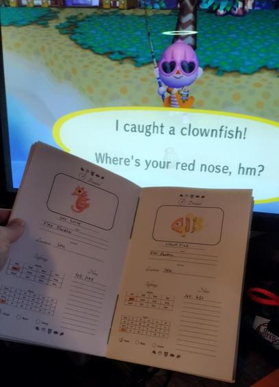 Photo of a screen playing City Folk. The player has caught a clown fish. In front of the screen, the player holds up the journal open to the clownfish page. There's a drawing and details have started to be filled in. Next to it is the page for the seahorse