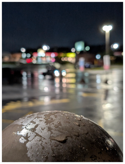 out of focus strip mall parking lot reflecting lights from vehicles and nearby business. in focus, in the foreground, the large round top of a brown plastic guard bollard is wet with rain.